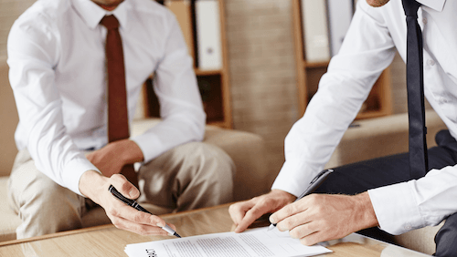 Kaksi kaulaspaitaan pukeutunutta miestä tutkii pöydällä olevaa neuvotteluesitystä.