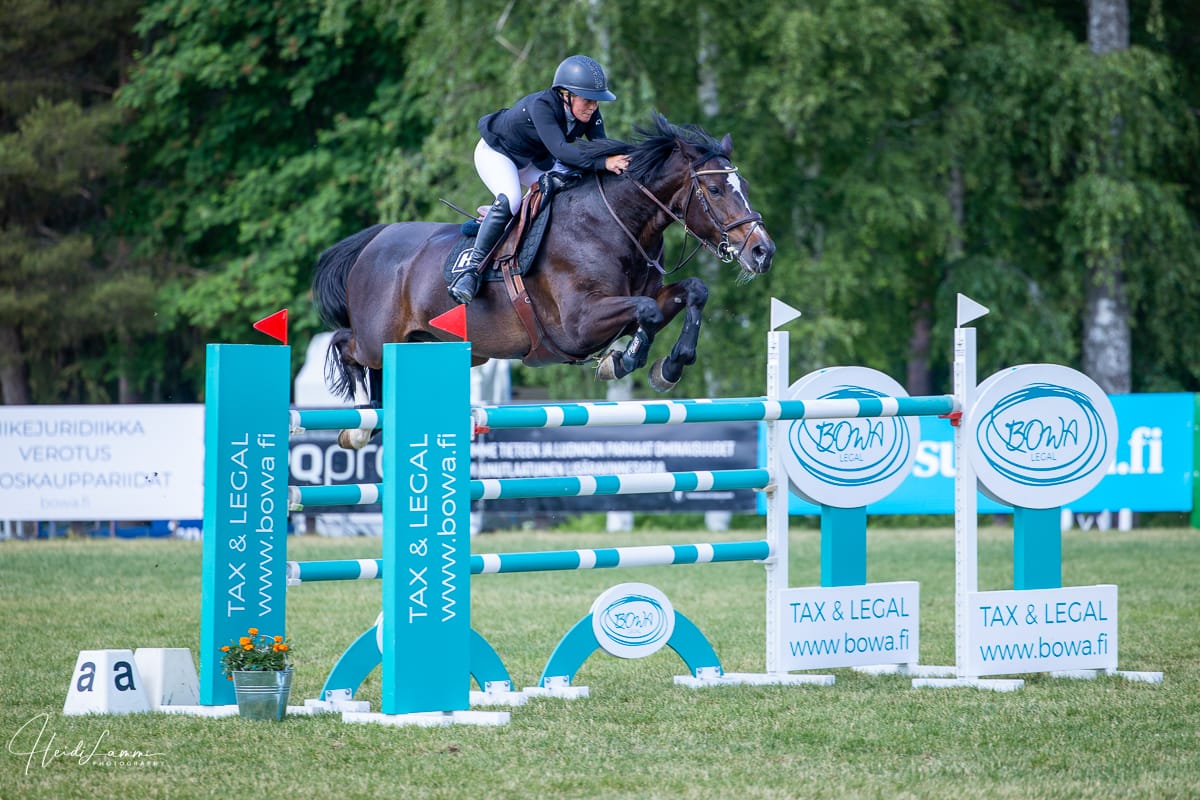 BOWA-este-c-HeidiLammi-10 • BOWA Legal Horse and rider mid-air clearing a turquoise-and-white show jump with sponsor banners in the background, green field behind.